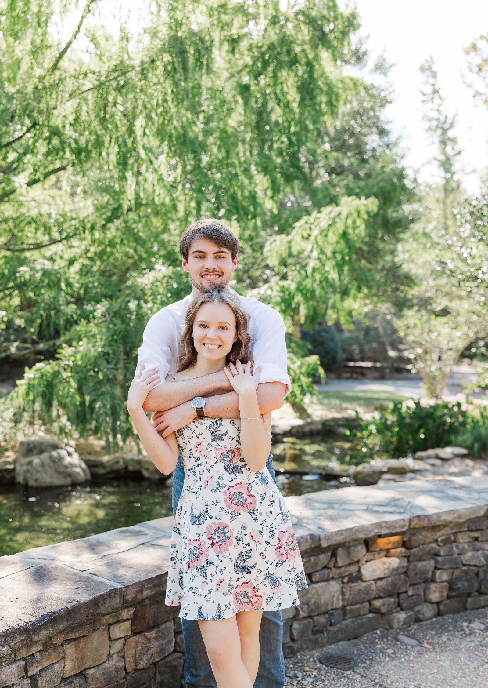 Engaged couple on a stone wall by the pond, framed by a weeping willow, at Shiloh Gardens