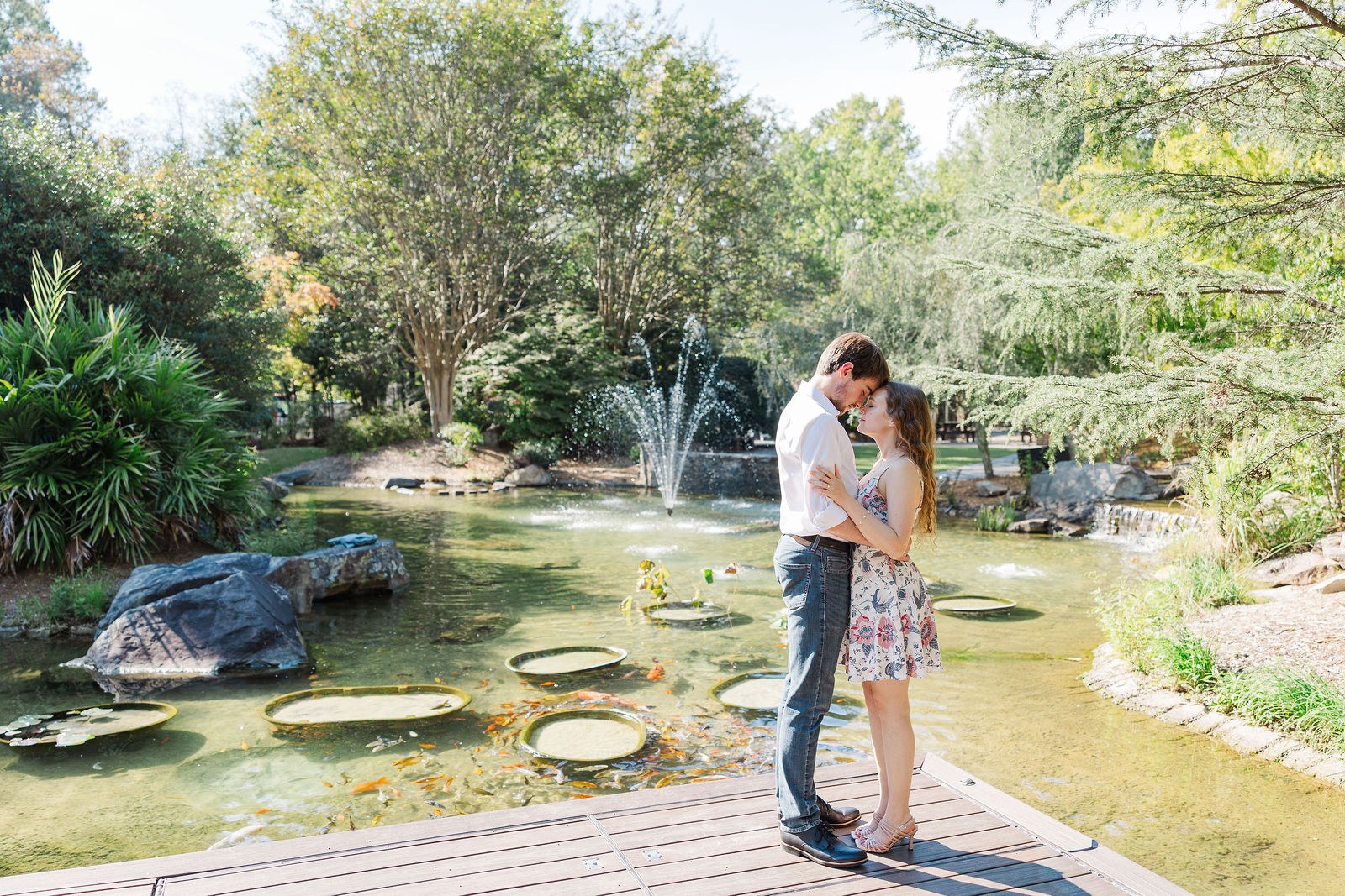 Dylan and Emma kiss on a deck over the lily pond with the fountain behind them at Shiloh Gardens