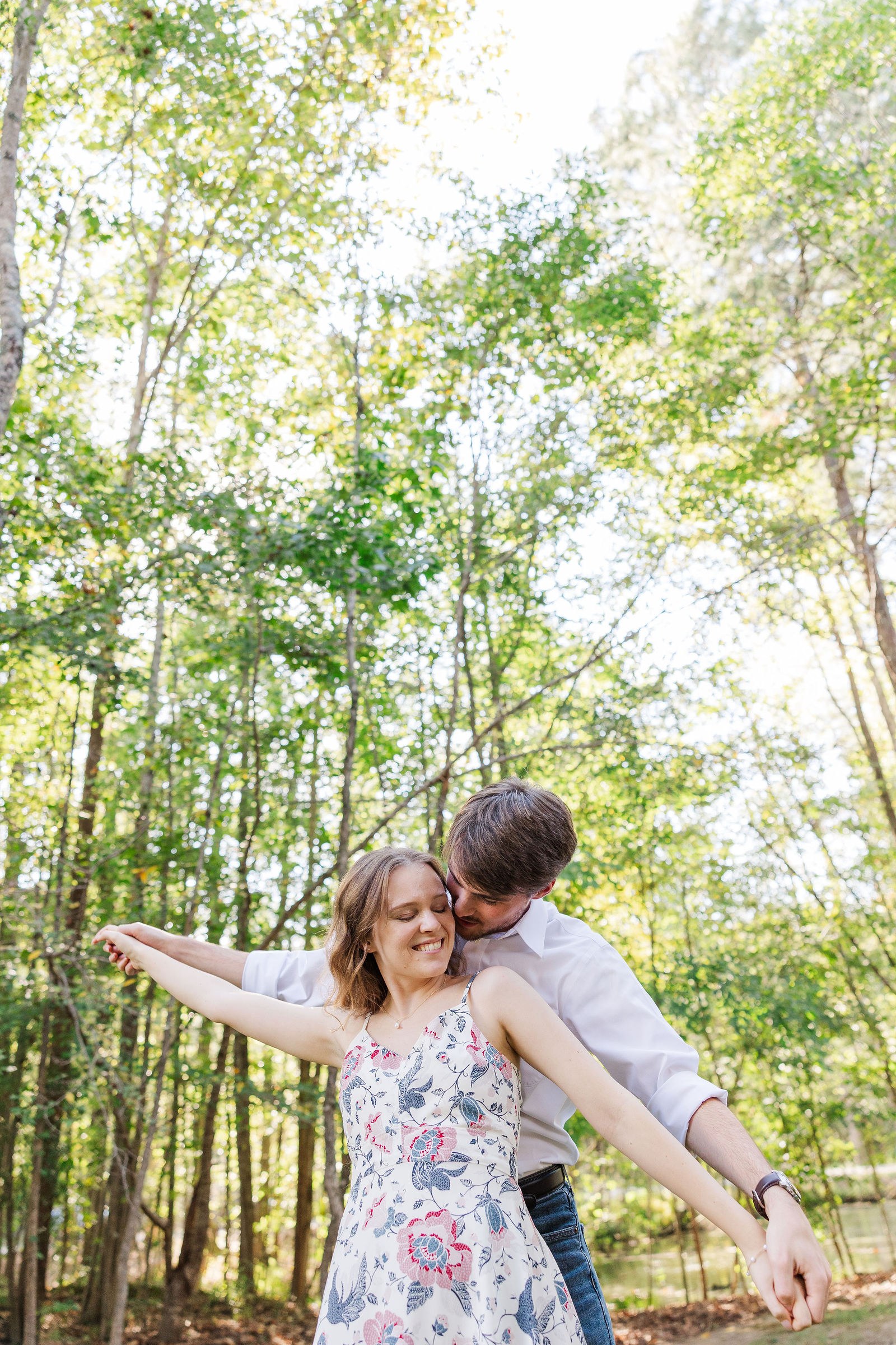 Emma laughs with her arms spread as Dylan holds her among tall trees in Cumming, Georgia
